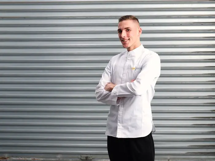 A male chef in a white coat stands with arms crossed in "Top Chef France".