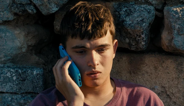 A teen talks on a blue phone by a stone wall in Enzo.