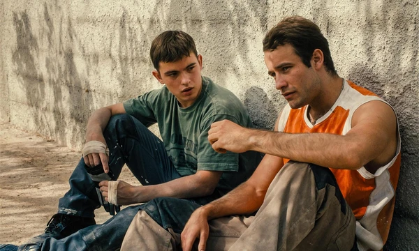 Two men converse while sitting against a wall in Enzo.